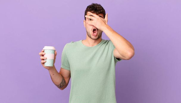 man with coffee looking shocked, scared or terrified, covering face with hand and peeking between fingers - Foto, Bild