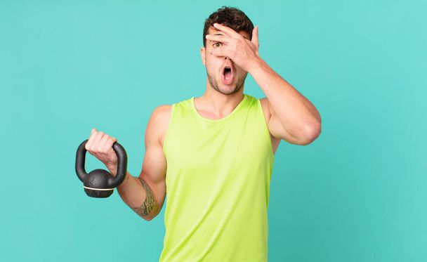 fitness man looking shocked, scared or terrified, covering face with hand and peeking between fingers - 写真・画像