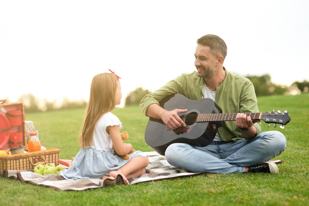 Cute little girl in casual clothes sitting on a green grass in park and looking at her loving dad playing guitar - Photo, Image