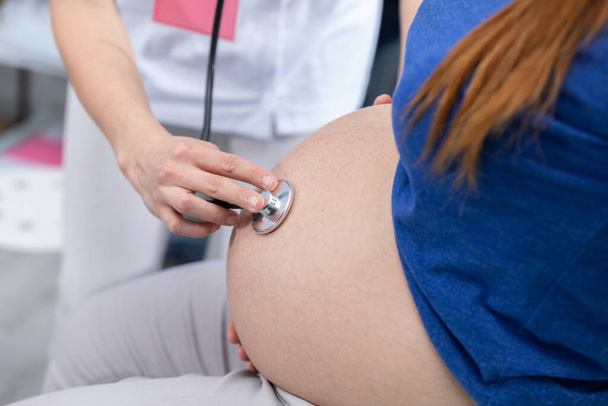 Close-up view of a lady doctor auscultating a pregnant womans abdomen with a stethoscope. Professional doctors office - Foto, immagini