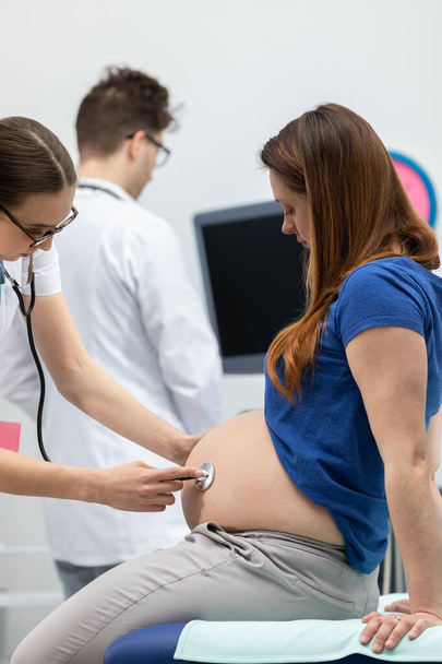 A lady doctor auscultates the abdomen of a pregnant woman with a stethoscope. In the background, a doctor is standing next to an ultrasound machine - Foto, imagen