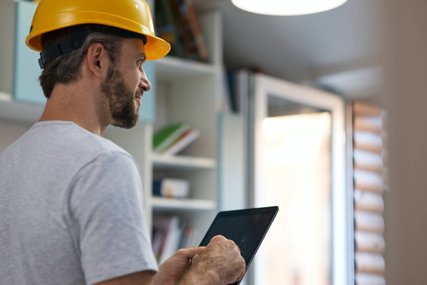 Professional repairman wearing helmet looking aside, using digital tablet while standing indoors - Foto, imagen