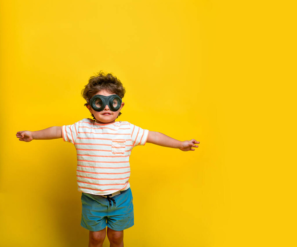 Funny smiling kid in aviator glasses and helmet isolated on yellow background. Child playing with a plane. Vintage pilot concept. Toys for kindergarten and preschool children - Photo, Image