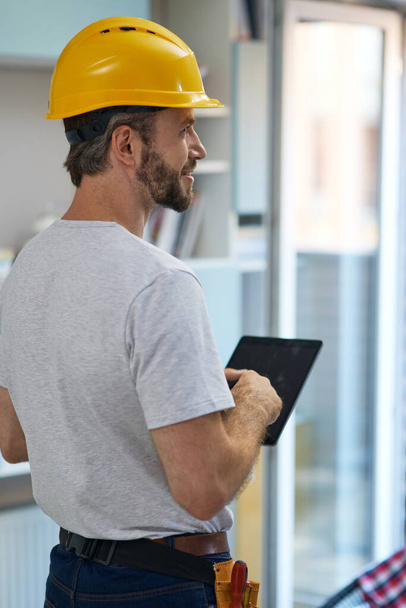 Reparador profesional con casco mirando a un lado, utilizando tableta digital mientras está de pie en el interior - Foto, Imagen