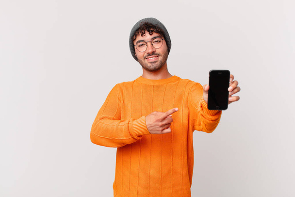 hispanic man with a cell smiling cheerfully, feeling happy and pointing to the side and upwards, showing object in copy space - Foto, Imagem