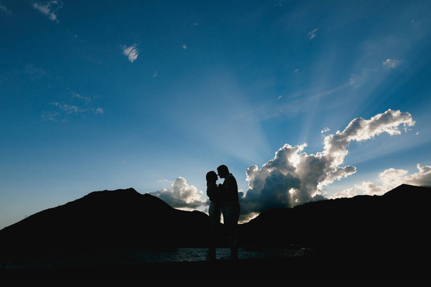 Silhuetas de um homem e uma mulher que se abraçam e se beijam ao pôr-do-sol na praia contra o fundo das montanhas  - Foto, Imagem