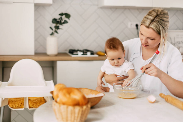 Молодая мать держит своего ребенка на руках, взбивает яйца венчиком и готовит тесто. Мама и ребенок проводят время вместе - Фото, изображение