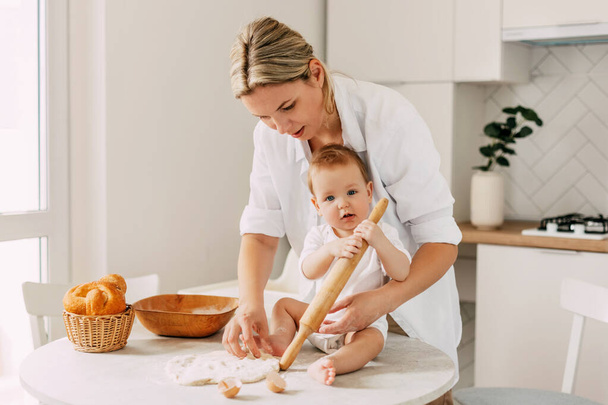 A cute baby holds a rolling pin in his hands, and his mother rolls out the dough for a pie. Time together. Childhood, motherhood, parenthood - Photo, Image