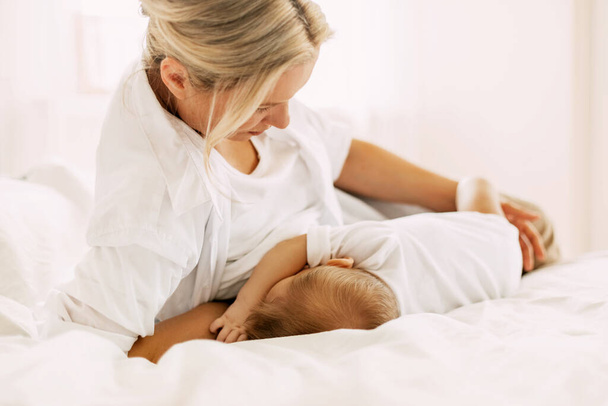 Eine junge Mutter stillt ihr Neugeborenes, während sie auf dem Bett liegt. Pflege, Kindheit, Mutterschaft - Foto, Bild