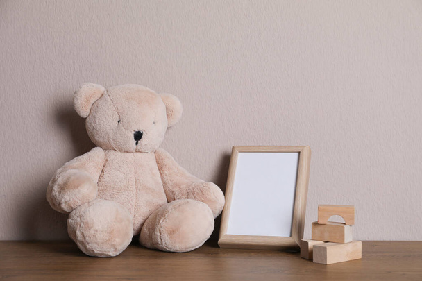 Empty photo frame, teddy bear and building blocks on wooden table near grey wall. Space for design - Foto, afbeelding