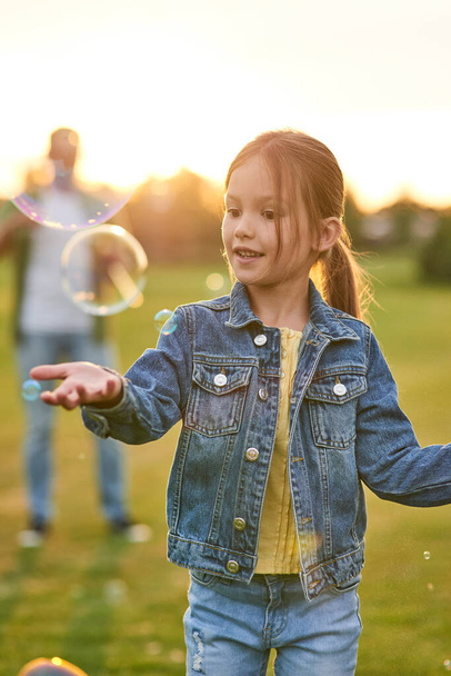 Прекрасна маленька дівчинка розважається з мильними бульбашками в сонячний день, дочка і тато проводить час разом на відкритому повітрі
 - Фото, зображення