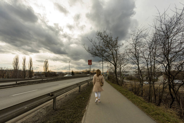 Krakow, Poland - April 01, 2021: Rear view of a female girl walking as pedestrian on a side walk alley next to busy highway of Cracow against dramatic clouds - Photo, Image