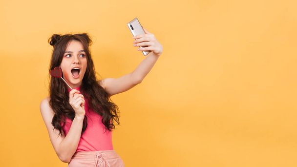 Una linda joven de color rosa sostiene una piruleta en forma de corazón en sus manos, sonríe y toma una selfie en su teléfono inteligente. Lugar para su texto - Foto, Imagen
