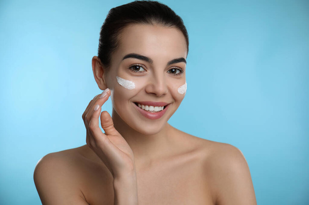 Young woman applying facial cream on light blue background - Photo, image