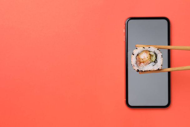 Holding tasty sushi roll with chopsticks over smartphone on red background, top view. Space for text - Foto, afbeelding