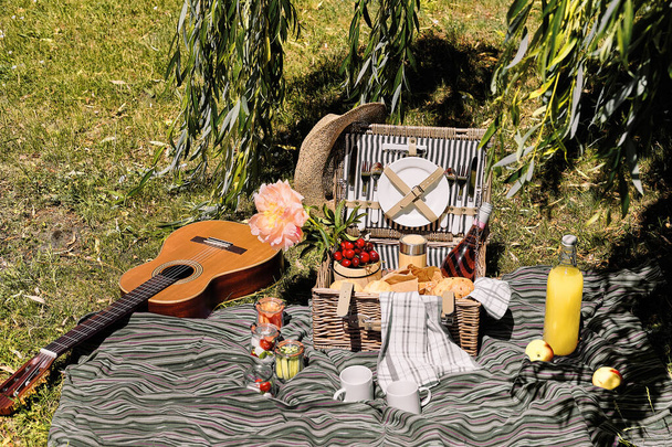 Vintage picnic basket, hamper with baguette and lemonade outdoors on a grass with cheese, mozzarella, tomatoes, cherries, vine. Guitar on straw blanket and stripy towels.Eco friendly picnic al fresco. - Photo, image