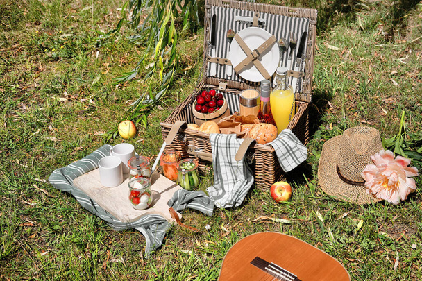 Vintage picnic basket, hamper with baguette and lemonade outdoors on a grass with cheese, mozzarella, tomatoes, cherries, vine. Guitar on straw blanket and stripy towels.Eco friendly picnic al fresco. - Fotó, kép
