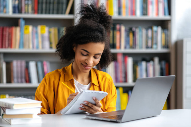 Vidám, okos, koncentrált afro-amerikai diáklány, jegyzetel egy jegyzetfüzetben, online előadásokat hallgat, ismerkedik, miközben ül az asztalnál az egyetemi könyvtárban, mosolyog - Fotó, kép