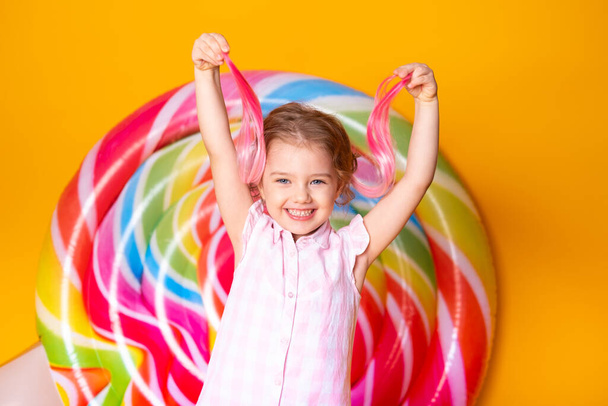 Happy cute little girl in pink dress and pink hair having fun on yellow background with lollipop. - Photo, Image
