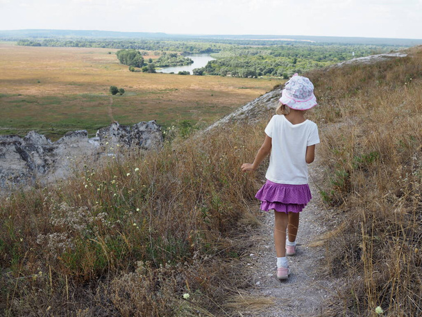 vista dalla parte posteriore una bambina cammina attraverso le colline di fronte a lei un bellissimo paesaggio con un fiume. - Foto, immagini