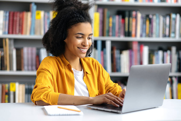 Glückliche attraktive afrikanisch-amerikanische Mädchen in stilvollen Kleidern, sitzt Studentin in der Universitätsbibliothek am Schreibtisch, bereitet sich auf die Prüfung oder Lektion, surft im Internet, sucht Informationen, lächelt - Foto, Bild