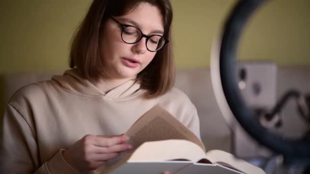 Chica blogger libro con gafas de ojo grabación vlog en cámara digital smartphone en casa. Mujer joven que usa luz de anillo para registrar su blog de libro, reseña de libro - Imágenes, Vídeo
