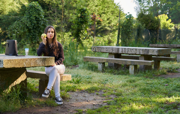 Die schöne junge Frau in schwarzem Hemd sitzt auf einem Steintisch und isst einen Apfel in der Natur mit ihrem Handy, einem Kaffee und ihrer Tasche mit Bäumen im Hintergrund. Veganer Teenager macht Pause im Park - Foto, Bild