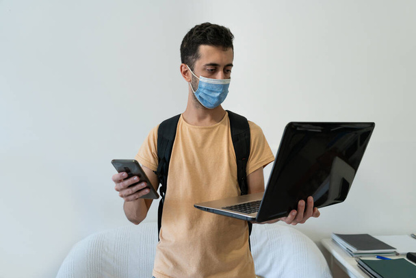 Φοιτητής σε απομόνωση στο σπίτι φορώντας μάσκα και κρατώντας ένα φορητό υπολογιστή και τηλέφωνο. Έννοια ασφάλειας για τη μελέτη. - Φωτογραφία, εικόνα