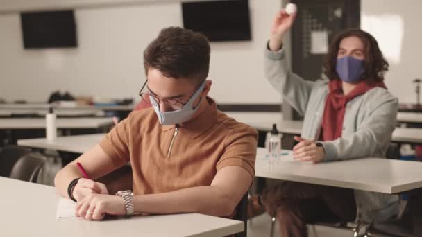 Średni długi męski student rasy kaukaskiej siedzący przy biurku, mający zajęcia, rzucający papierową piłkę w kolegę z klasy mieszanej przed sobą, przyjaciel odwracający się i pokazujący kukułcze gesty. - Materiał filmowy, wideo