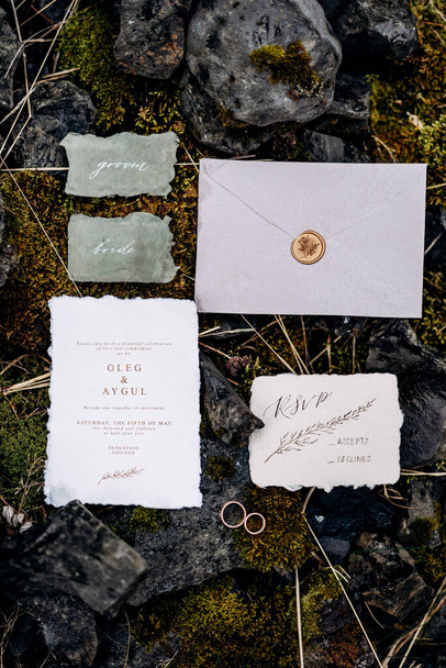 Wedding rings lie on moss-covered stones, surrounded by decorated invitations and envelope sealed with sealing wax. Nearby are two signs with inscriptions. Inscription: groom, bride - Photo, Image