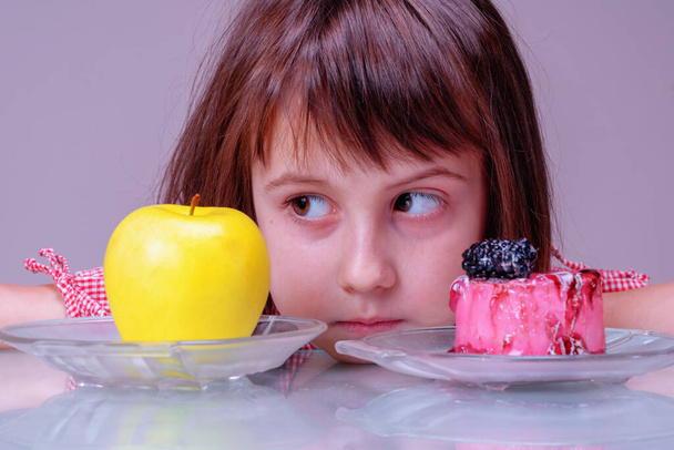 Ernährungs- und Ernährungskonzept. Schwierige Wahl zwischen Obst und Süßigkeiten. Porträt eines jungen schönen Mädchens, das zwischen Apfel und leckerem Kuchen wählen kann. Horizontales Bild. - Foto, Bild