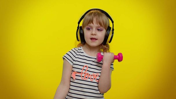 Niña escuchando música a través de auriculares, ejercitando los músculos del brazo levantando pesas rosadas - Foto, imagen