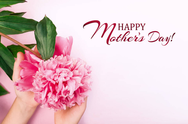 Woman hold in hands a pink peony, leaves on a white background. Fresh flowers. Inscription Happy Mother's Day. Advertising banner, poster for Mother's Day. Flat lay, top view, close up, copy space - Photo, Image