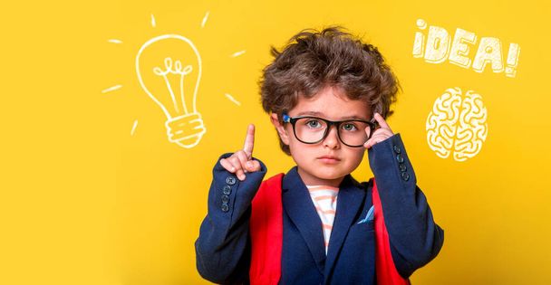 Small smart child in glasses with bag isolated on yellow. Little genius student thinking with lightbulb and math formulas. Student has idea. Pupil with picture of brain. Kindergarten kid go to school - Photo, Image