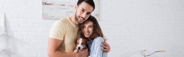 cheerful multiethnic couple hugging with jack russell terrier in modern living room, banner - Foto, imagen