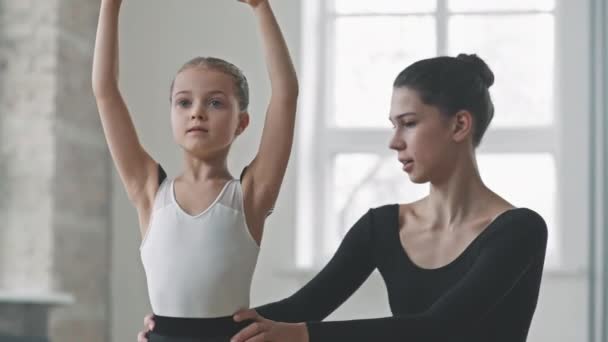 Keskikokoinen laukaus söpö 9-vuotias tyttö oppimisen baletti aiheuttaa ja liikkeitä naisten kouluttaja auttaa häntä, käyttäessään kirkas parvi-tyylinen tanssistudio - Materiaali, video