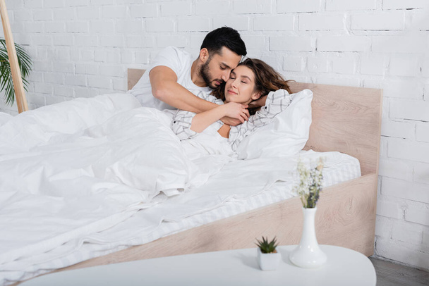 smiling multiethnic couple with closed eyes hugging and lying on bed - Foto, Bild