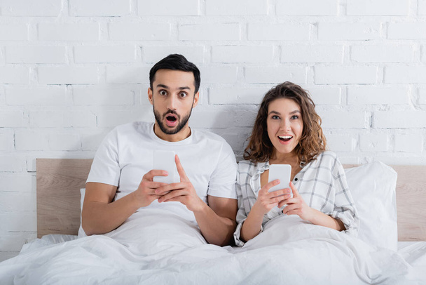 изумленная межрасовая пара лежит на кровати и использует смартфоны  - Фото, изображение