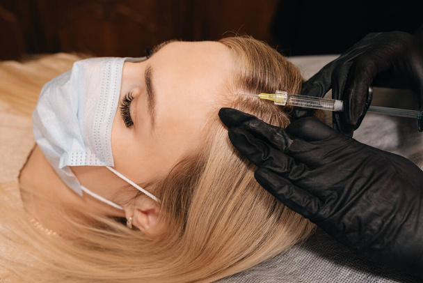 Kosmetikerin in Handschuhen gibt Injektion in den Kopf der blonden Frau. Mesotherapie, Haarausfall-Therapie. - Foto, Bild