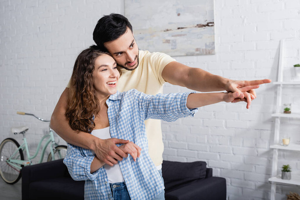 happy multiethnic man and woman hugging and pointing with fingers at home - Photo, image
