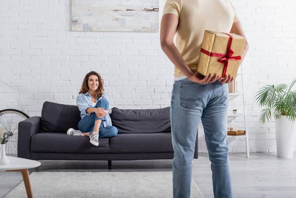 blurred man hiding present behind back near happy woman in sofa  - Photo, Image