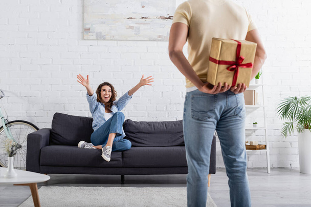blurred man hiding present behind back near excited woman in sofa  - Photo, Image
