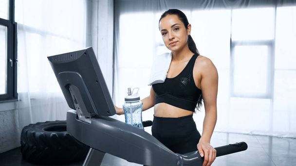 femme sportive en sueur avec serviette sur tapis roulant regardant la caméra près de la bouteille de sport avec de l'eau dans la salle de gym - Photo, image