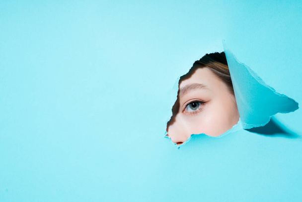 Painted eye and eyebrow of a young girl looking at the camera through a hole in a blue background - Photo, image