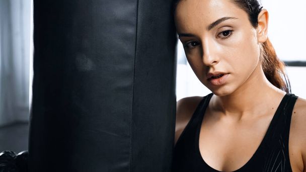 tired young sportswoman leaning on punching bag in gym  - Photo, Image