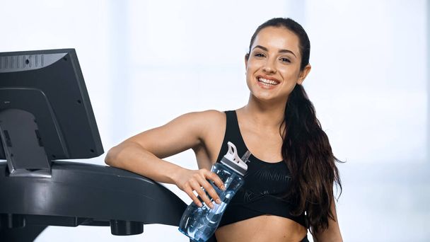happy woman in sportswear holding sports bottle with water near treadmill  - 写真・画像