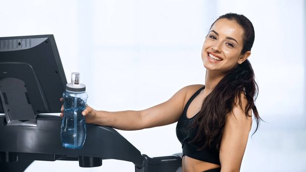 smiling woman in sportswear holding sports bottle with water near treadmill  - Photo, Image