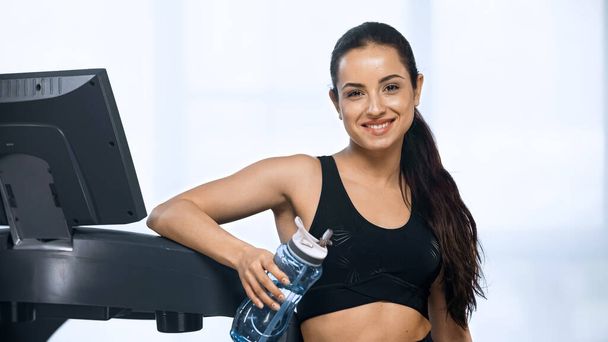 cheerful woman in sportswear holding sports bottle with water near treadmill  - 写真・画像