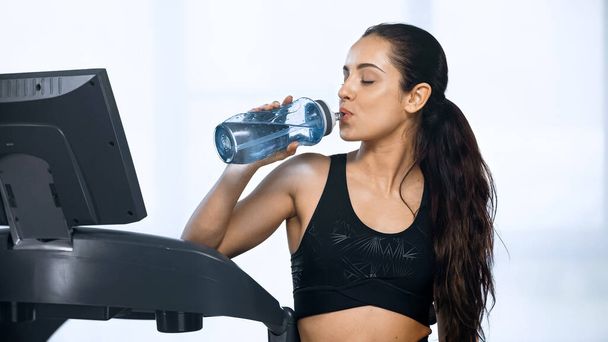 young woman in sportswear holding sports bottle and drinking water near treadmill  - 写真・画像