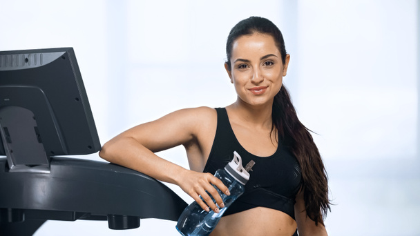 young pleased woman in sportswear holding sports bottle with water near treadmill  - Фото, зображення